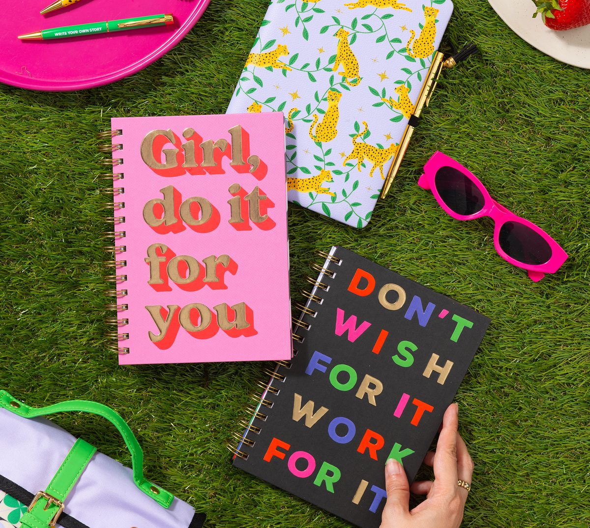 Three notebooks with bold lettered and cheetah-print covers, styled on grass with pens and sunglasses