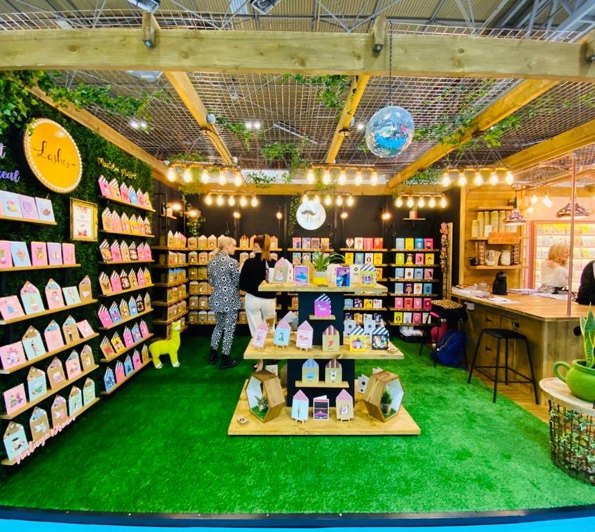 Tache’s trade-show stand seen from the front, with card displays on a grass-green floor under a timber frame and string lights