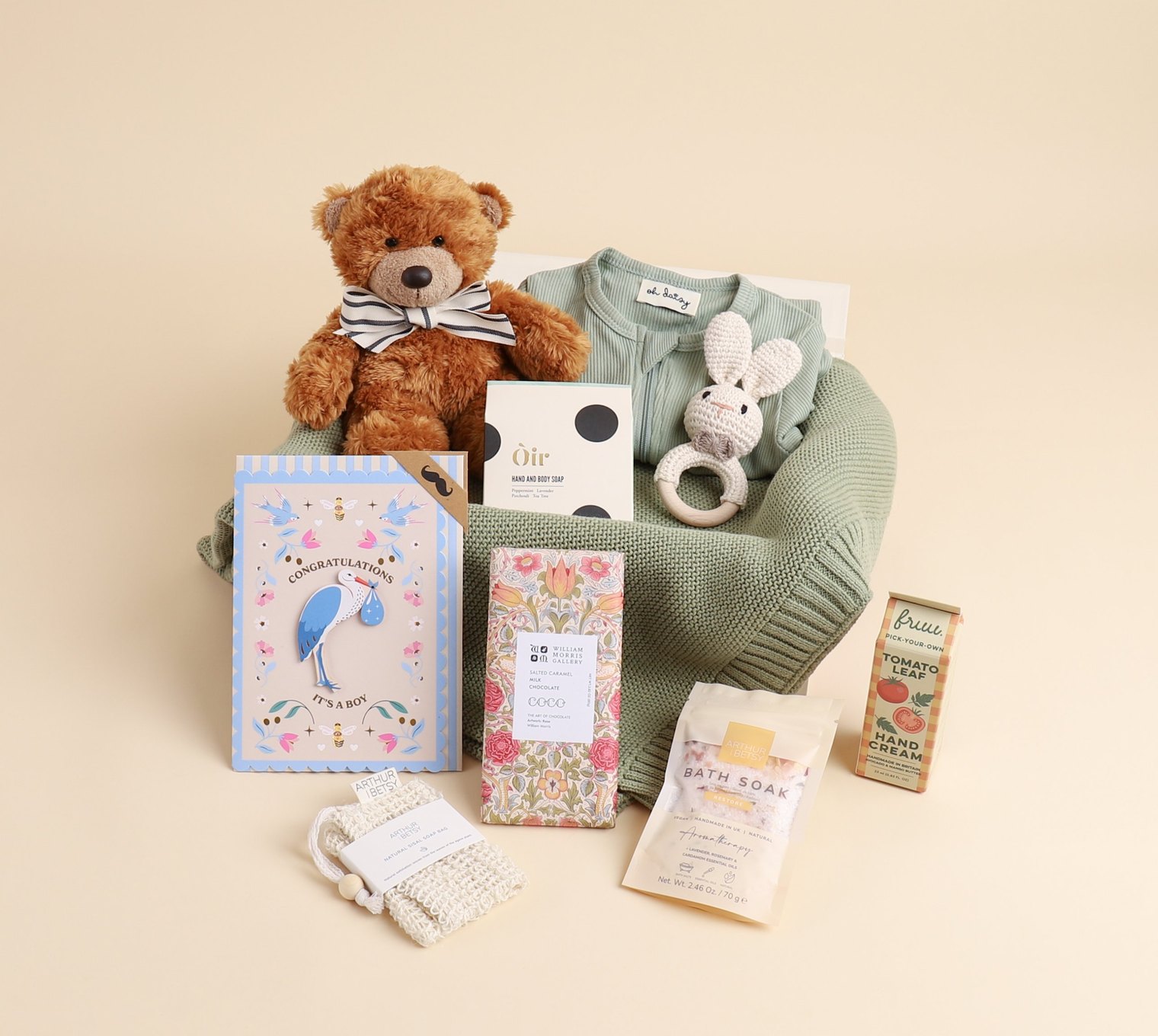 An Oh Daisy baby hamper on a cream backdrop: a teddy bear, a knitted green blanket, a sleepsuit with the Oh Daisy label, a crocheted bunny rattle and a Tache “It’s a boy” card