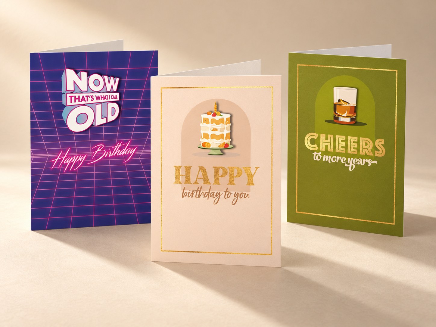 Three greeting cards standing together: a neon “Now that’s what I call old” birthday card, a gold-foil “Happy birthday to you” cake card and a green “Cheers to more years” card